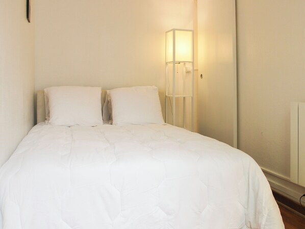 Una cama bien hecha con ropa blanca en una habitación con pared blanca y una lámpara de noche.