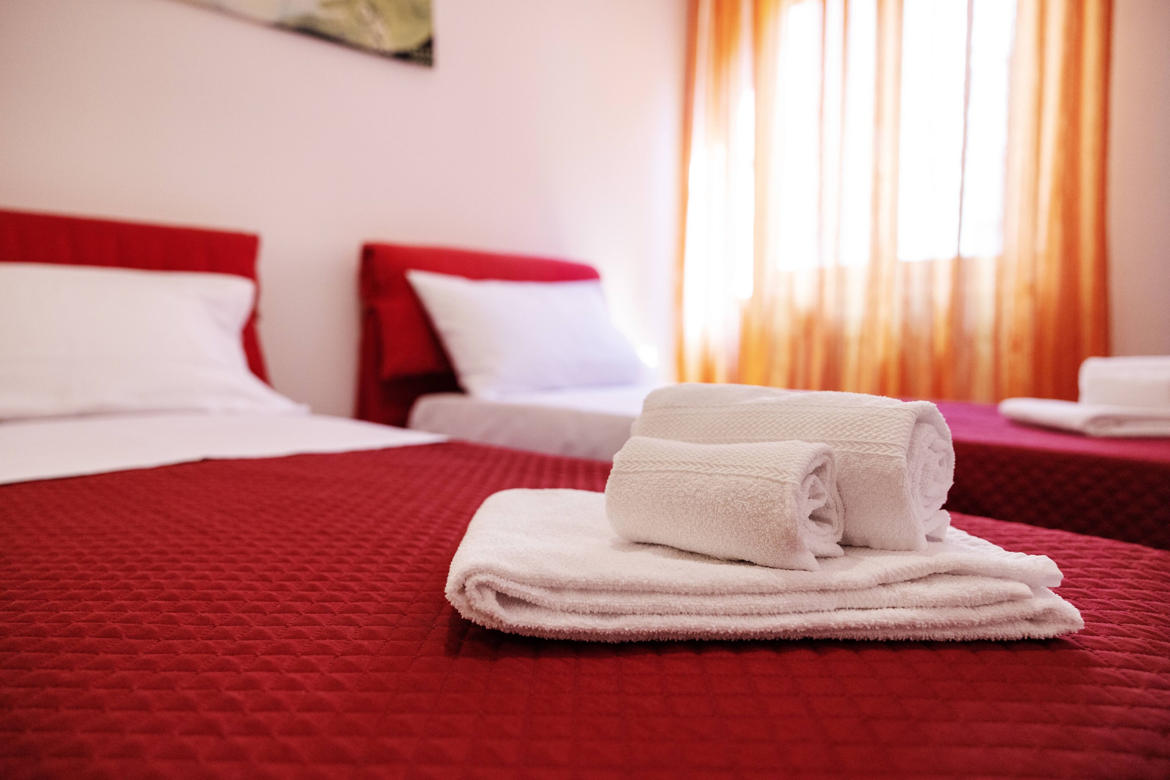 Une chambre d’hôtel avec deux lits, une literie rouge, des oreillers blancs et une serviette blanche soigneusement pliée posée sur l’un des lits.