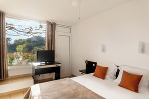 A hotel room with a bed, a desk with a television, a window with curtains, and a view of greenery outside. - Apartment for 2–4 people by the sea in Cargèse (Cargèse)