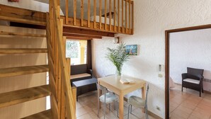 Una habitación con escalera de madera, una mesa de comedor con sillas, una planta en maceta y un cuadro en la pared.
