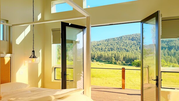 Un dormitorio con cama, lámpara colgante y vistas a una ladera boscosa a través de puertas abiertas.