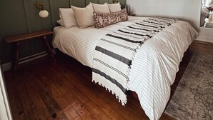 Een slaapkamer met houten vloer, een bed met gestreept beddengoed, een bankje en twee ramen met uitzicht op bomen.
