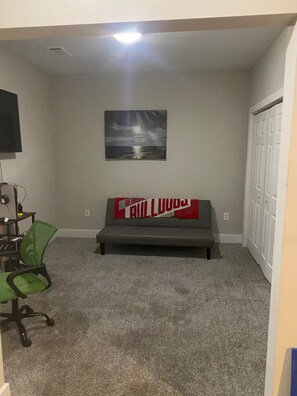 Une chambre avec un canapé gris, une couverture rouge et blanche, un bureau avec un ordinateur et une chaise.