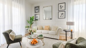 Ein Wohnzimmer mit einer Couch, Sesseln, einem Couchtisch mit Früchten, einem Beistelltisch mit einer Lampe und gerahmten Bildern an der Wand.