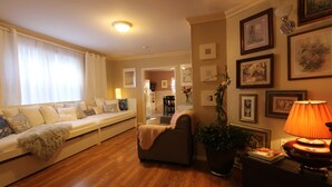 Ein Wohnzimmer mit Sofa, Sessel und einem Tisch mit Lampe. An der Wand hängen gerahmte Bilder und es steht ein Blumentopf bereit.