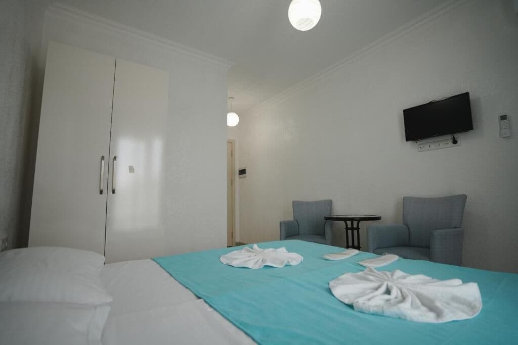 Quarto de hotel com uma cama, duas poltronas, uma mesa pequena, uma televisão e um guarda-roupa.