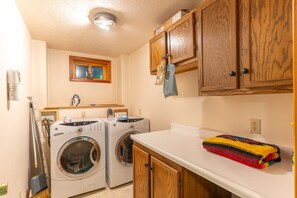 A laundry area with two front-loading washing machines, wooden cabinets, a countertop, and a window above the washing machines. - Whitefish Mountain Farmhouse | Near Glacier (Whitefish)