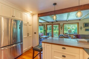 A kitchen with a stainless steel refrigerator, wooden cabinets, a dining area with a table and chairs, and a view of the outdoors through a door. - Whitefish Mountain Farmhouse | Near Glacier (Whitefish)