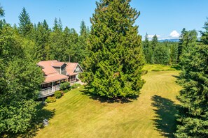 A large house with a porch, surrounded by a well-maintained lawn and dense forest. - Whitefish Mountain Farmhouse | Near Glacier (Whitefish)
