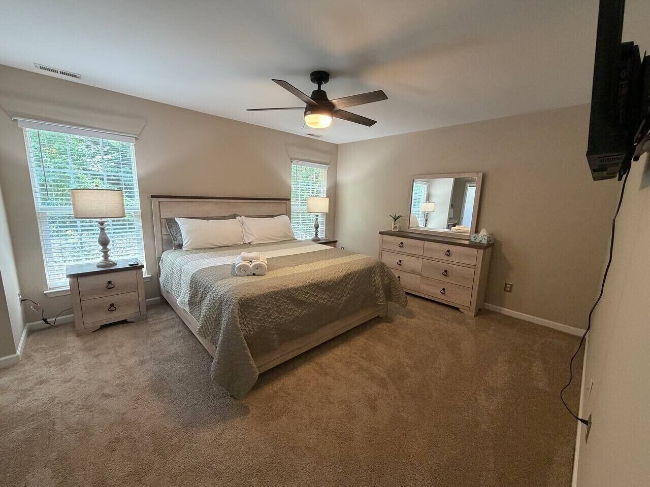 Ein Schlafzimmer mit Bett, Nachttischen, Kommode, Deckenventilator und Fenstern.