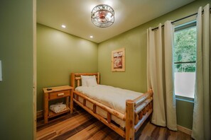 A single bed with a wooden frame, a bedside table with a drawer, a hanging light fixture, a window with curtains, and a framed picture on the wall.