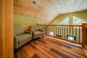 A loft-style room with a sloped ceiling, wooden flooring, two sofas, a wooden railing, and a large window.