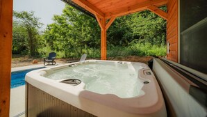 A hot tub in a wooden gazebo with a pool and chairs outside.
