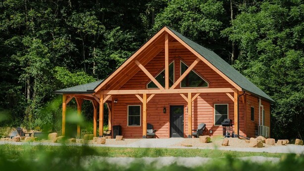 A log cabin with a gabled roof, a porch with seating, and a door leading to the interior.