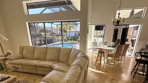 Uma sala de estar espaçosa com um amplo sofá de couro bege em formato de L, uma área de jantar com mesa e cadeiras, e vista para uma piscina através de grandes janelas.