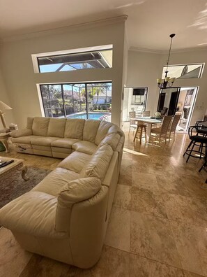 Ein geräumiges Wohnzimmer mit einer großen, L-förmigen beigefarbenen Ledersofa, einem Essbereich mit Tisch und Stühlen sowie Blick auf einen Swimmingpool durch große Fenster.