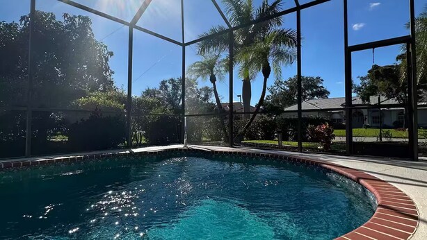 Uma piscina azul límpida com borda de tijolos, cercada por uma área coberta com estrutura metálica, palmeiras e céu claro.
