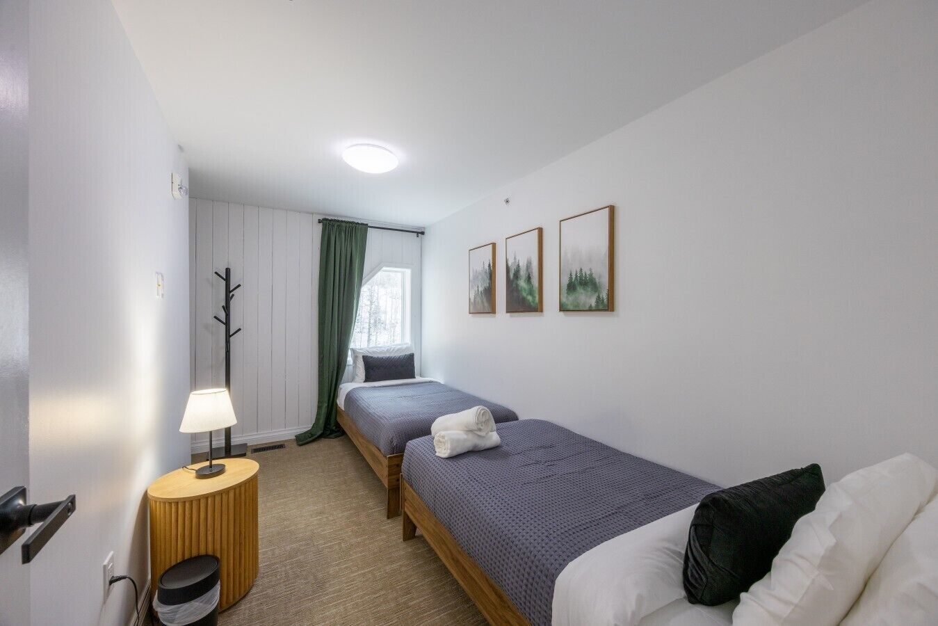 A hotel room with two beds, a bedside table with a lamp, a coat rack, and a window with curtains.