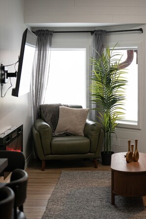 A compact living space with a green armchair, a large potted plant, a wooden side table, and a window with curtains.