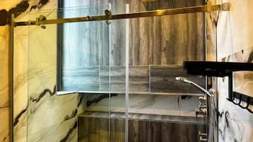 A modern bathroom with a glass shower enclosure, marble walls, and a wooden floor.
