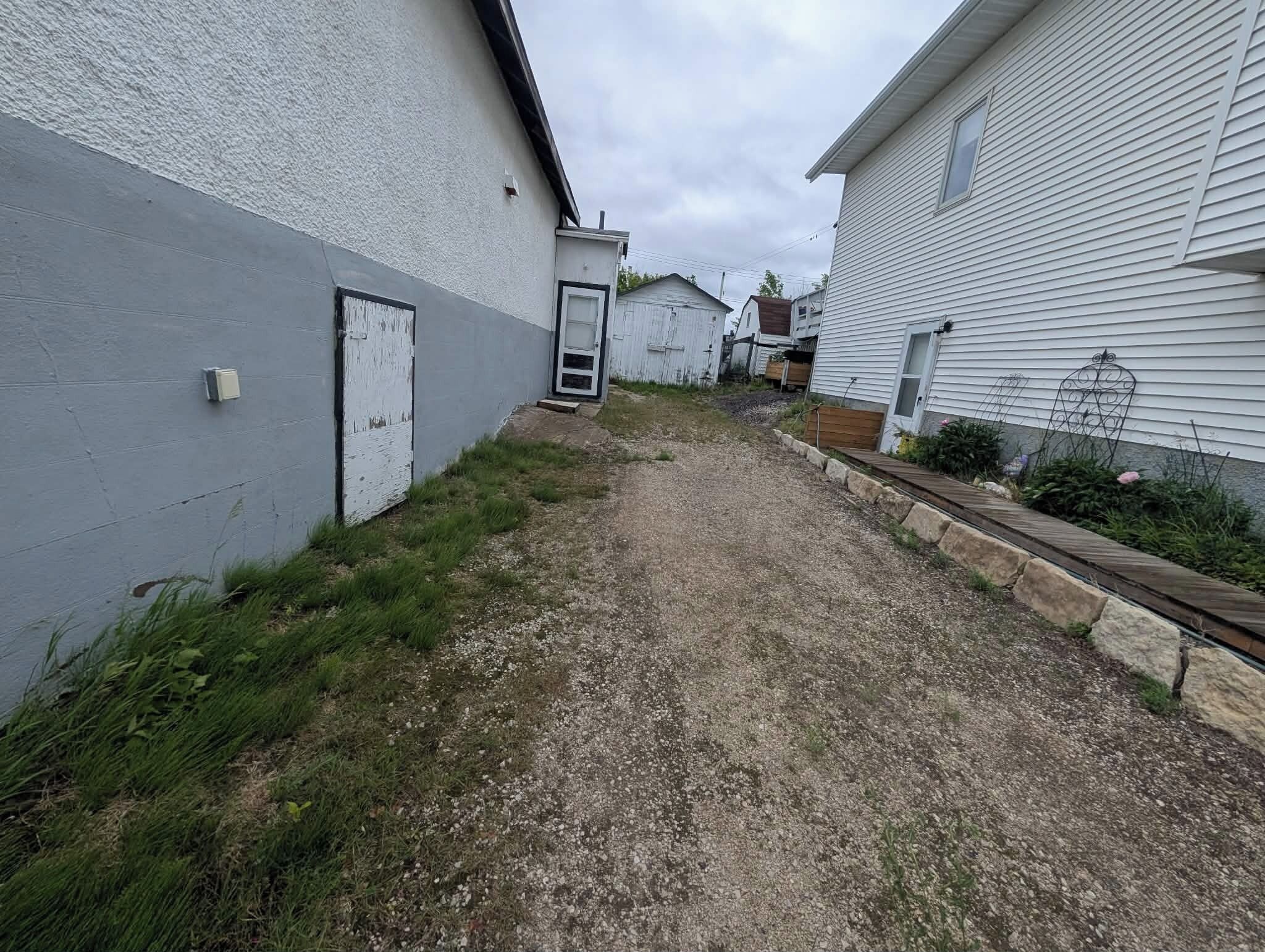A narrow gravel path between two white buildings with a metal gate and a small air conditioning unit.