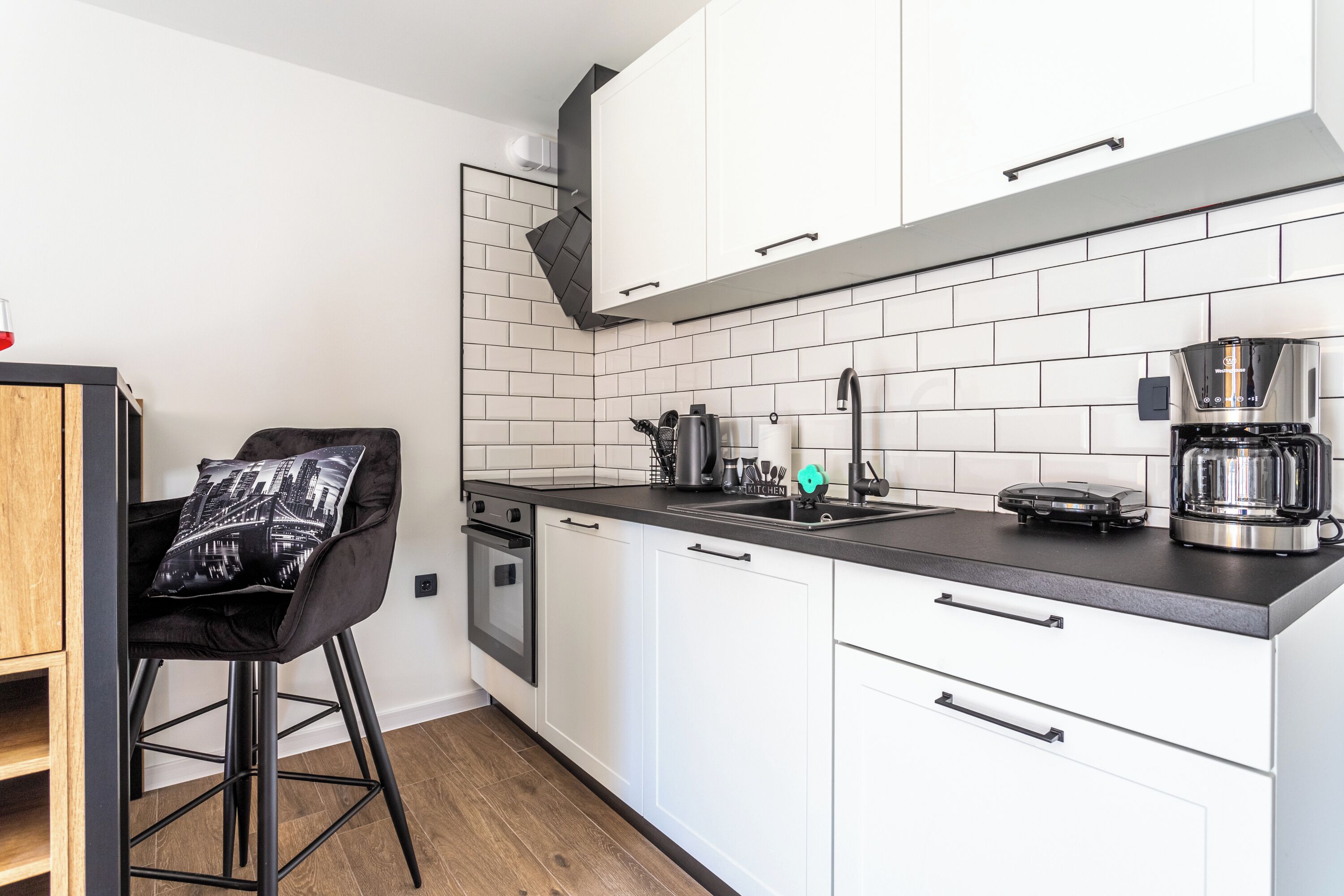A modern kitchen with white cabinets, a black countertop, a coffee maker, and a black chair with a cityscape cushion.