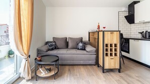 Un espacio habitable compacto con un sofá gris, una mesita redonda pequeña, un mueble de madera y una zona de cocina con armarios blancos y una campana extractora negra.