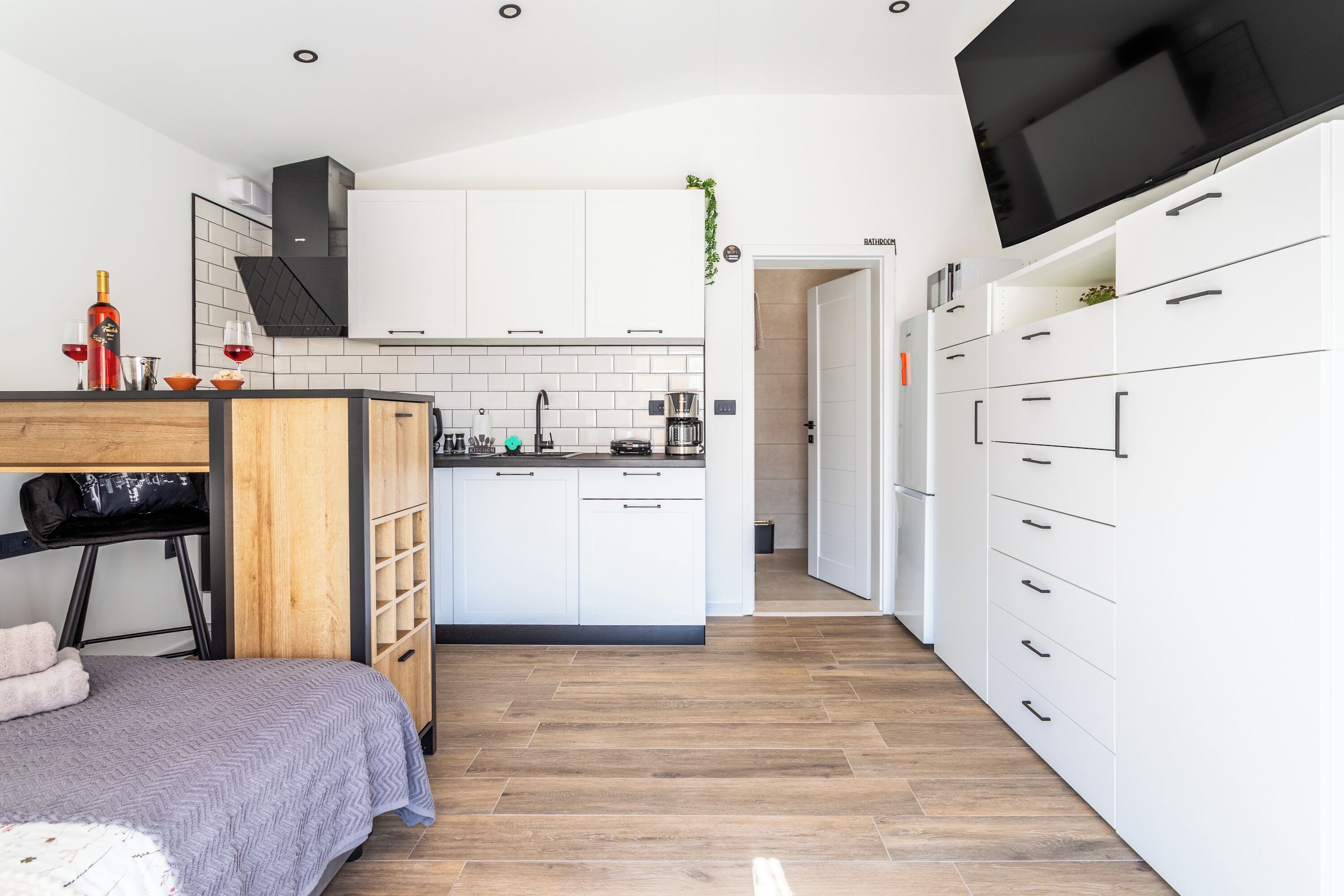 A modern kitchen with white cabinets, a wooden countertop, and a black TV mounted on the wall.
