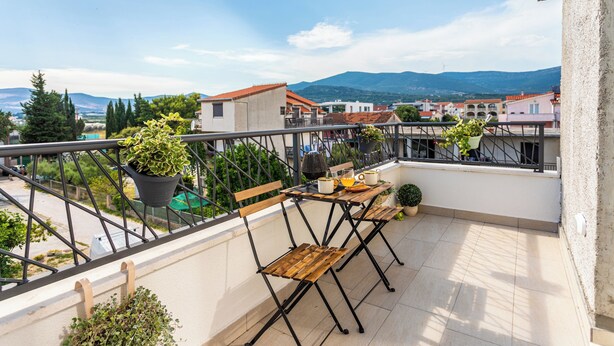 Un balcón con una mesa, sillas, plantas en macetas y vistas a edificios y montañas.