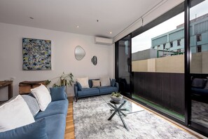 A modern living room with a blue sofa, a glass coffee table, and a large window offering a view of a building. - Stylish 3BDR Unit in the Heart of Melbourne CBD (Melbourne)
