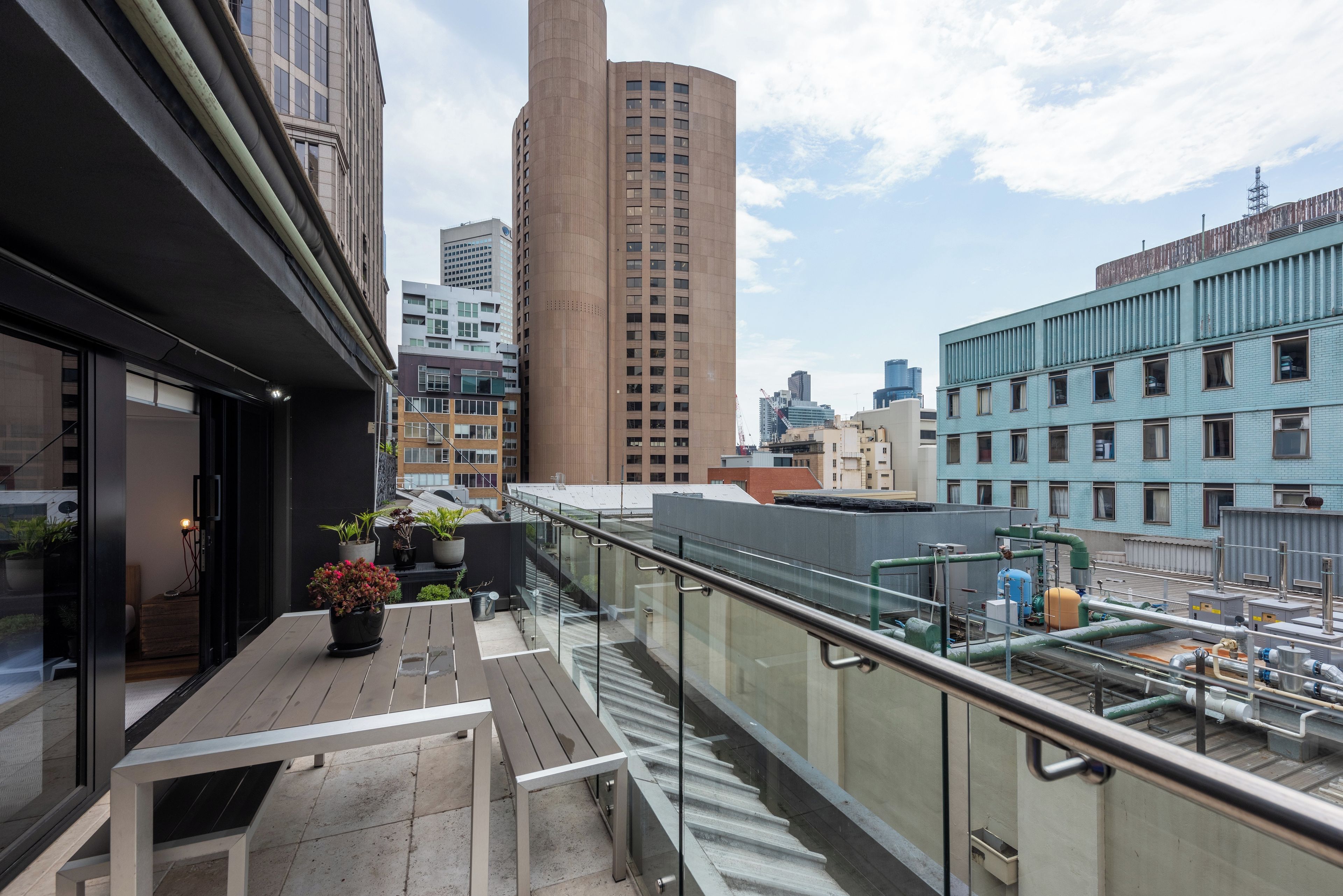 A rooftop terrace with a glass balustrade, potted plants, and a view of surrounding buildings.
