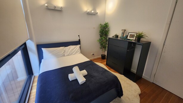 Un dormitorio con una cama, mesitas de noche, un aparador, una planta en maceta y un ventanal.