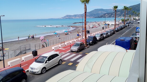 Un carrer costaner amb cotxes aparcats, una vorera i una platja al fons.