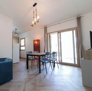 A modern living room with a dining table, chairs, a sofa, a TV, and a large window with curtains. - Villa Su Entu | with Garden close to the beach (Lanusei)