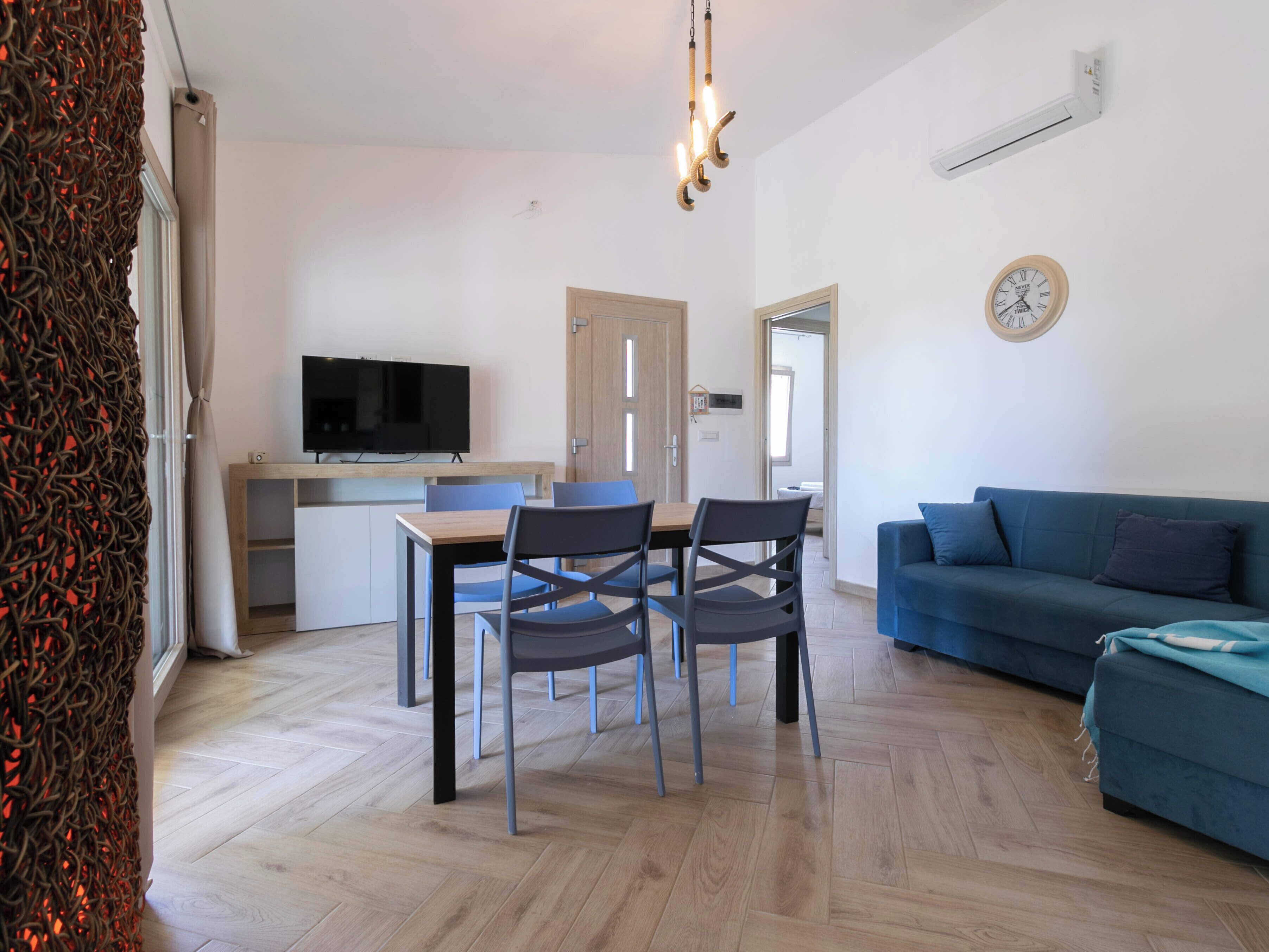 A modern living room with a blue sofa, a dining area with a TV and chairs, and a kitchen in the background.