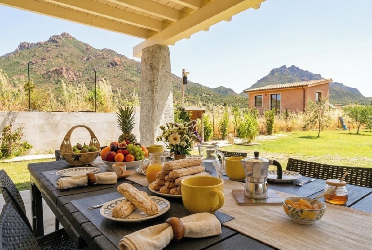 Une table de petit-déjeuner installée à l’extérieur, avec des fruits, des viennoiseries et du café, offrant une vue sur un paysage montagneux.