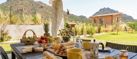 A breakfast table set outdoors with fruits, pastries, and coffee, overlooking a mountainous landscape.