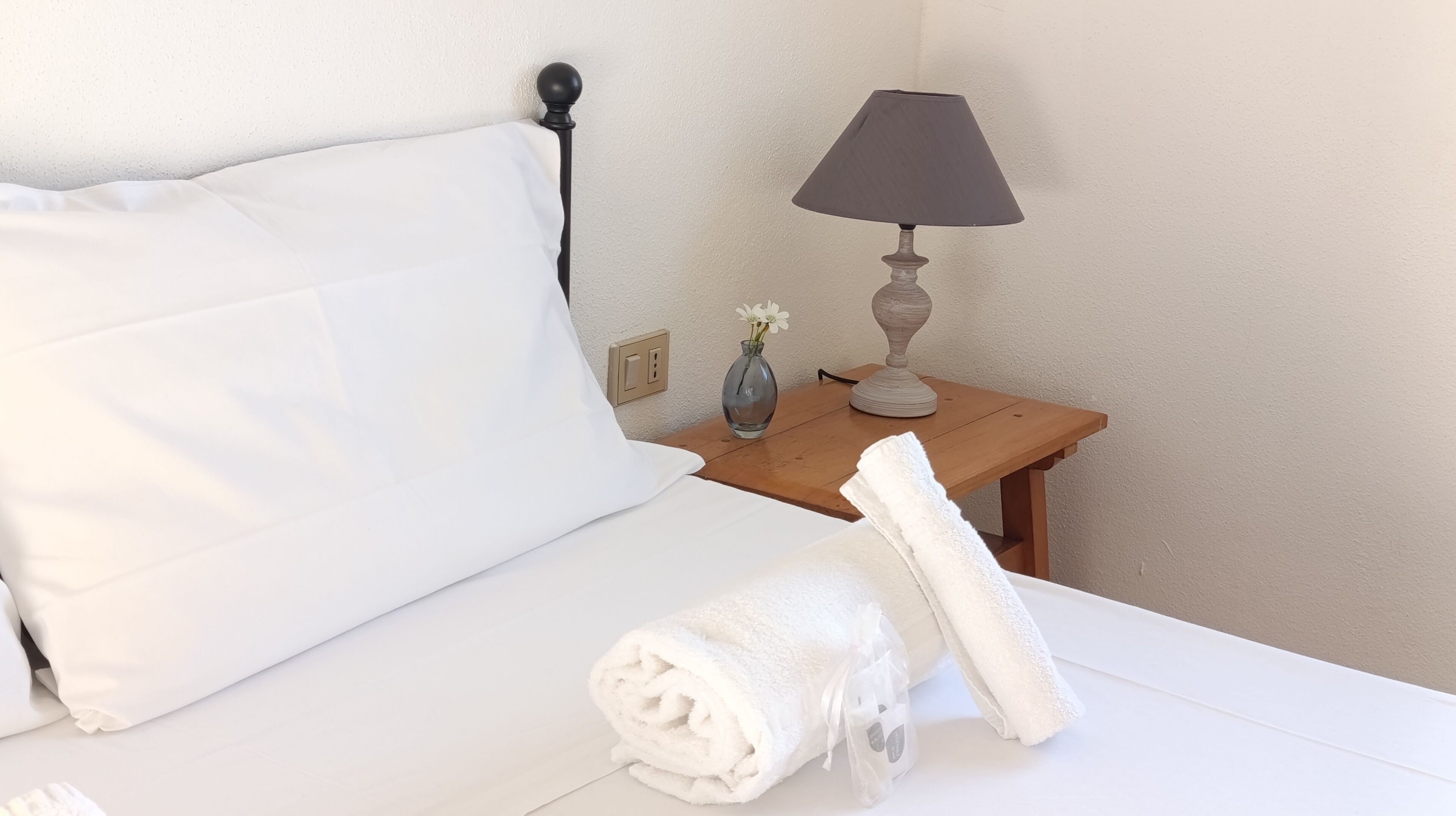Ein ordentlich gemachtes Bett mit weißen Leinentüchern, ein Nachttisch aus Holz mit einer Lampe und eine Vase mit Blumen.