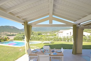 Un espace extérieur abrité où l’on peut prendre ses repas, avec une piscine et des montagnes en arrière-plan.