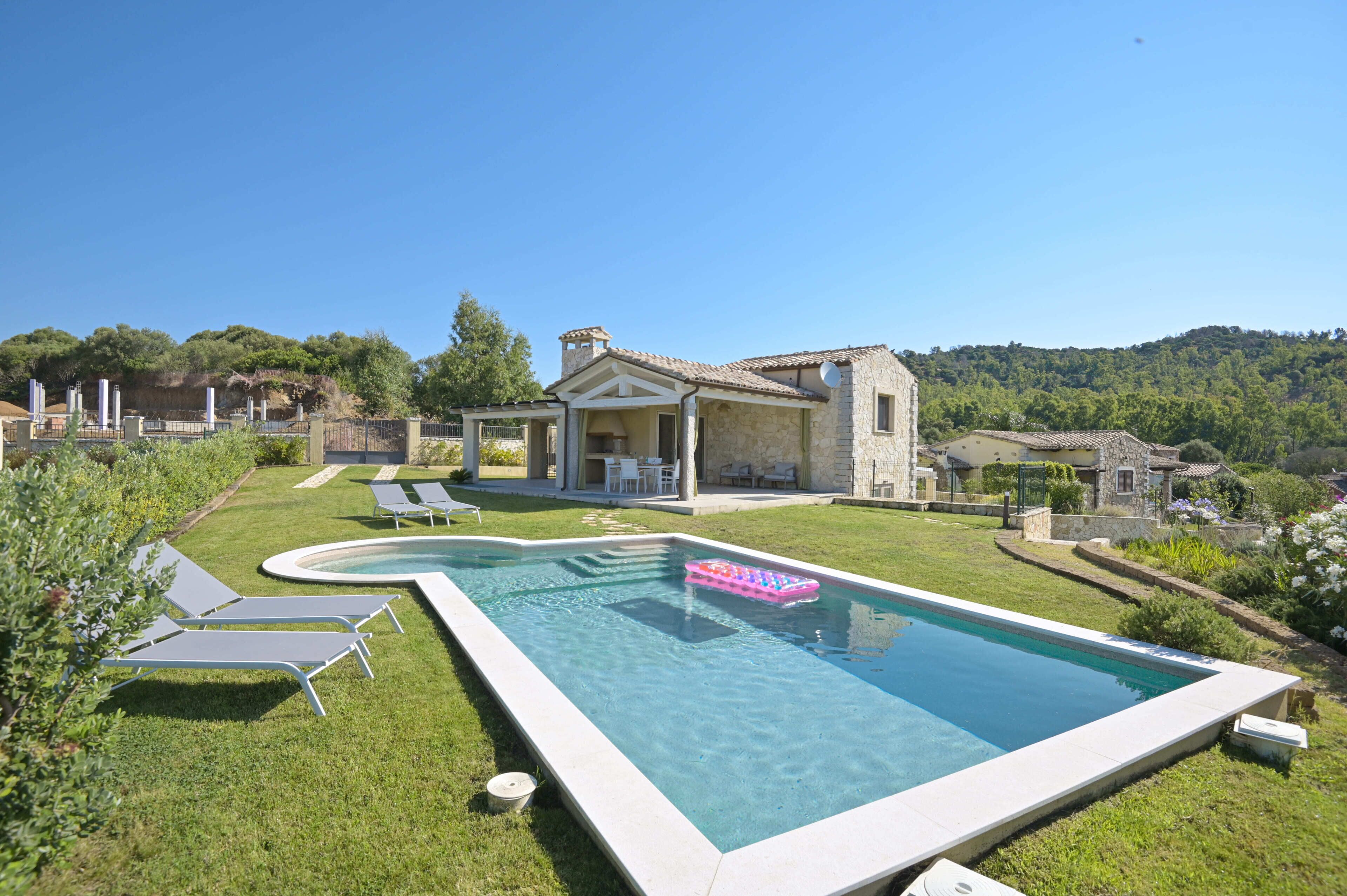A swimming pool with lounge chairs, a stone house, and a hilly landscape.