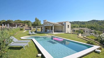A swimming pool with lounge chairs, a stone house, and a hilly landscape.