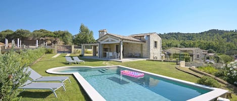 A swimming pool with lounge chairs, a stone house, and a hilly landscape.