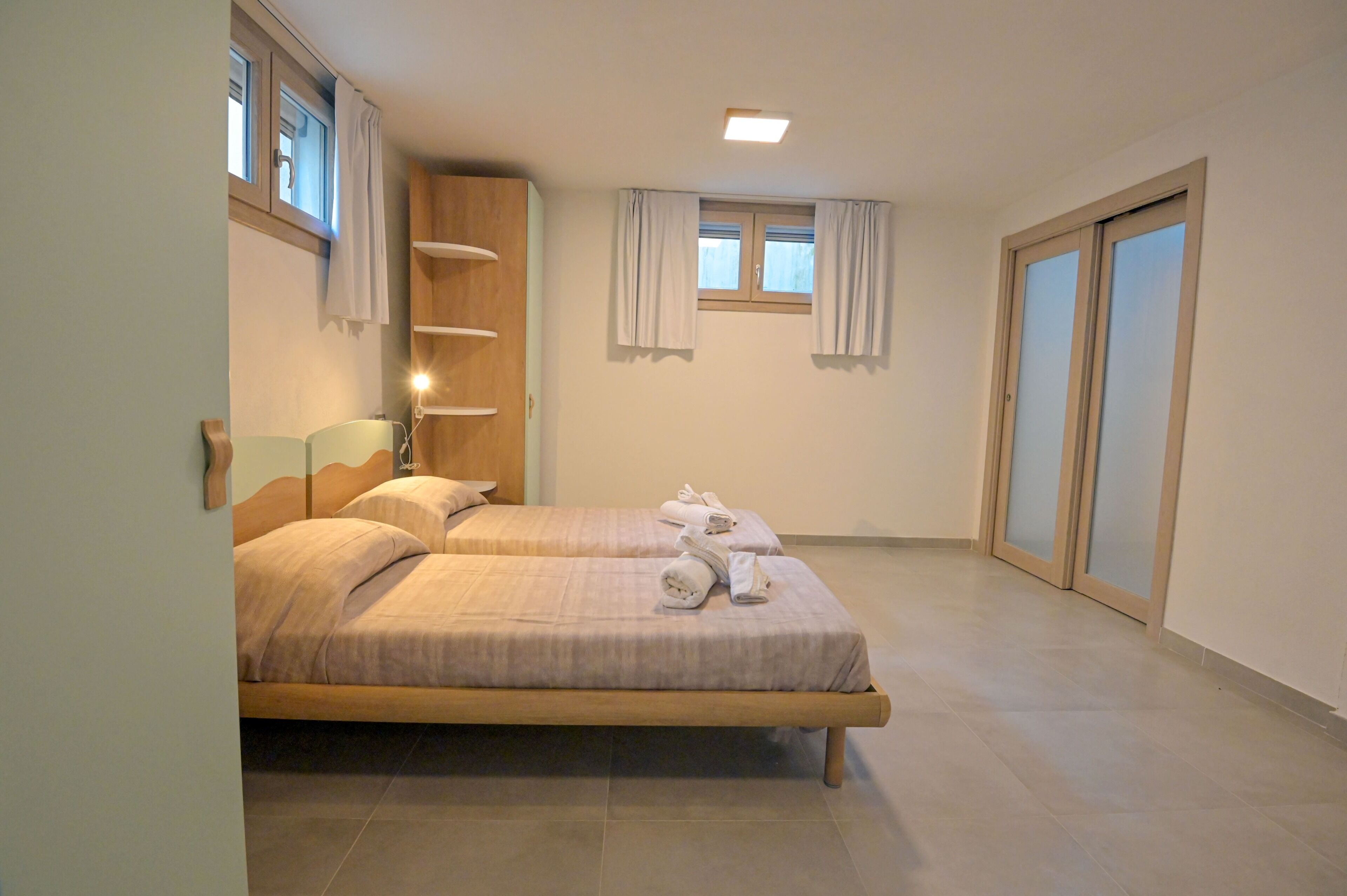 Habitación de hotel con dos camas, cabecero de madera, ventana con cortinas y puerta corrediza.