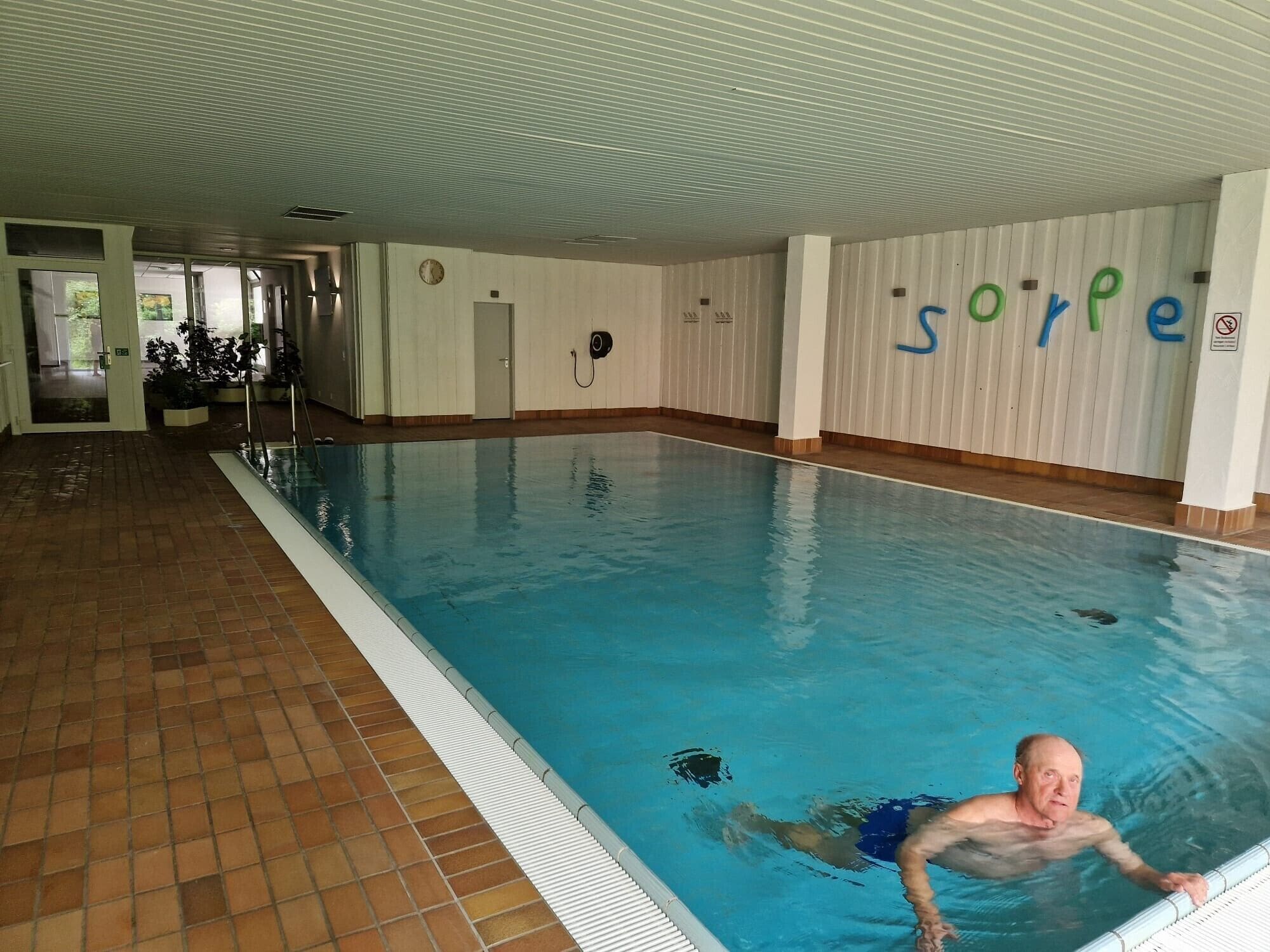 An indoor swimming pool with a tiled floor, a person swimming, and a wall with the word 'sorpe' visible.