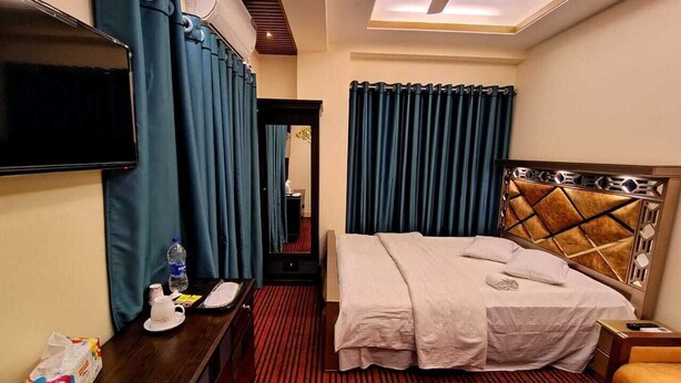 A hotel room with a bed, a desk with a flat-screen TV, a bedside table with a bottle and glasses, and a ceiling fan.