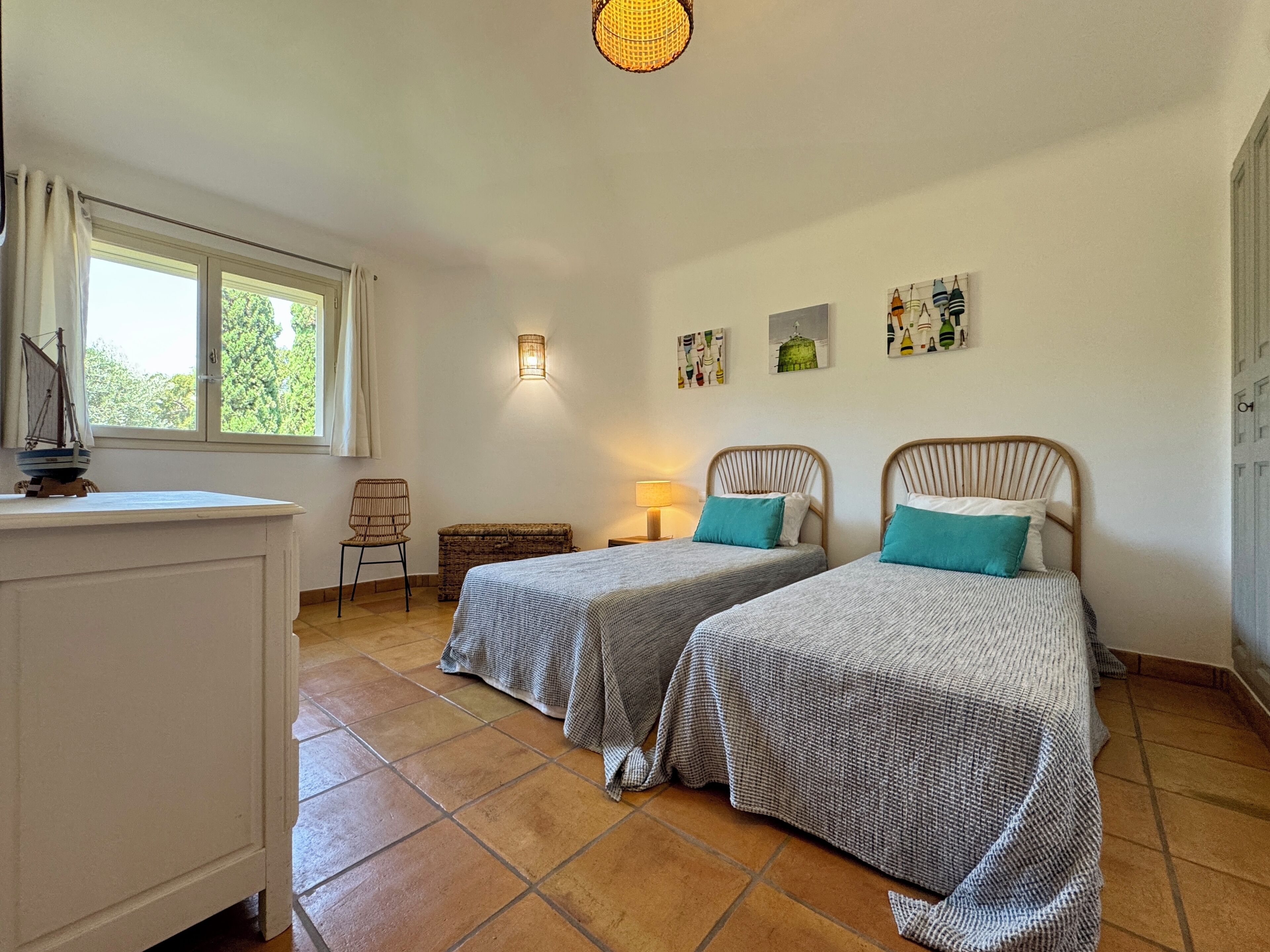 A bedroom with two beds, a window, a chair, a lamp, and artwork on the wall.