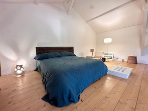 Ein Schlafzimmer mit Holzboden, einem Bett mit dunkelblauer Bettdecke, einem Nachttisch mit Lampe und einem Korbwannen-Korb.