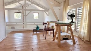 Ett rum med ett sluttande tak, trägolv, ett bord med stolar, en fläkt och fönster med gardiner.