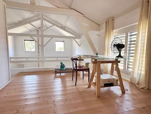 Ein Zimmer mit schrägen Wänden, Holzboden, einem Tisch mit Stühlen, einem Ventilator und Fenstern mit Vorhängen.