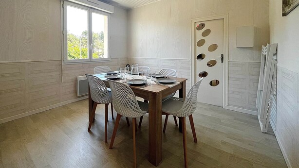 A dining area with a wooden table, white chairs, a red pendant light, and a window with a view. - RIVÉA - 2-bedroom apartment near the ocean (Mimizan)